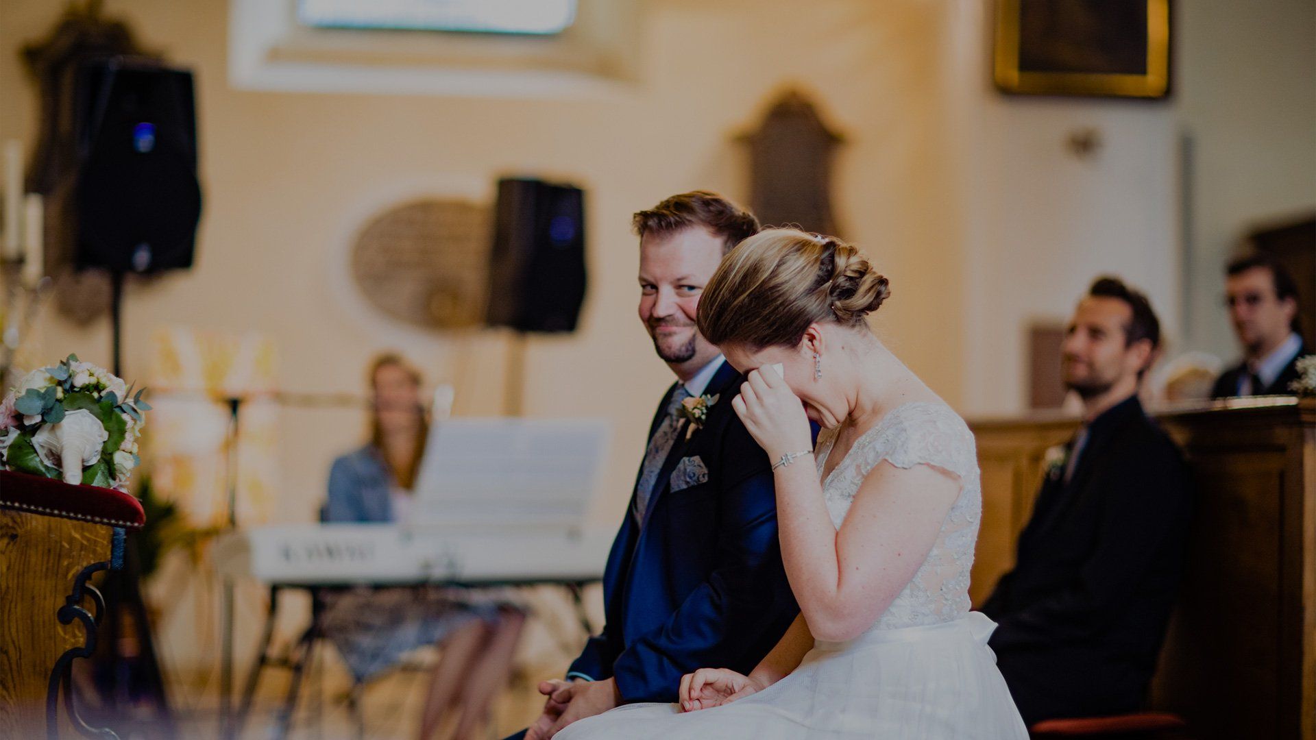 A bride and groom sat in a church, the bride is wiping her eyes.